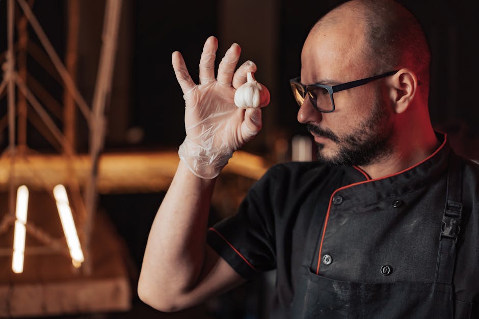 Bald chef wearing glasses holds garlic with gloves in a kitchen setting.