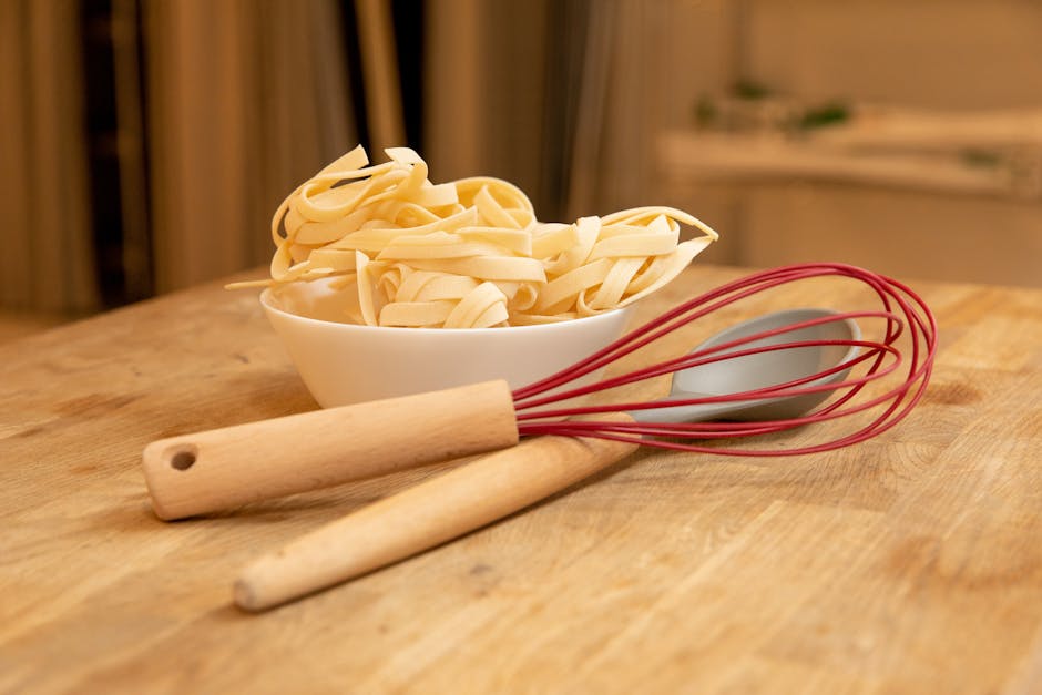 Wooden kitchen table with uncooked pasta in a bowl and utensils, ideal for cooking themed visuals.